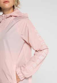 Chaqueta con capucha de color rosa claro, hecha de un material liso y resistente al agua. Presenta puños con elástico y texto de logo blanco a lo largo de la manga.
