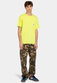 Neon yellow cotton t-shirt with pocket; paired with brown camouflage pants featuring leaf patterns. Footwear: blue athletic shoes.