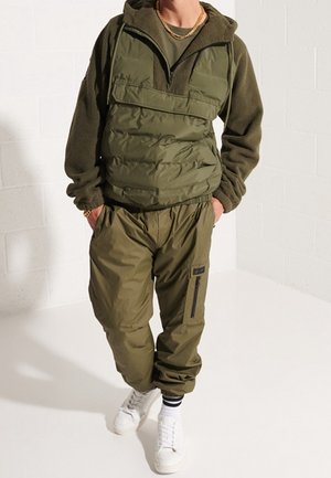 Olivově zelená prošívaná vesta se zipovým límcem a fleecovými rukávy, doplněná o kalhoty se stejným odstínem a elastickým pasem s kapsou na zip. Bílé tenisky.