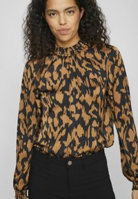 Blouse en imprimé léopard marron et noir avec un col froncé, des manches longues et une texture lisse, assortie à un pantalon noir.
