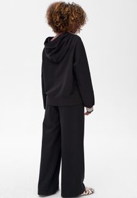 Czarna bluza z kapturem z opadającymi ramionami, w zestawie z szerokimi czarnymi spodniami. Posiada paski na mankietach. Gładka, miękka faktura.