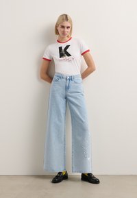 Weißes T-Shirt mit rotem Kragen und Logo-Grafik. Hellblaue weite Jeans mit gesticktem Text. Schwarze Schuhe mit gelben Socken.