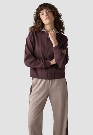 Woman with curly hair wearing a zipped dark brown jacket and beige pants with dark side panels against a plain background.