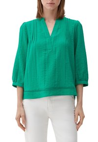 Grüne, strukturiert Bluse mit V-Ausschnitt, dreiviertellangen Ärmeln und horizontalem Paneel-Detail am Saum, kombiniert mit weißen Hosen.