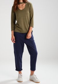 Olijfgroen, langarm T-shirt met een ronde hals en uitsnijdetails, gecombineerd met marineblauwe tapered broek en witte sneakers.