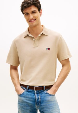 Giovane uomo sorridente che indossa una polo beige con piccolo logo sul petto, jeans blu e cintura in pelle marrone, mani nelle tasche.