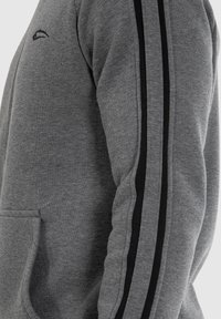 Sweatshirt gris avec des rayures noires sur les manches, présentant une poche avant et un logo subtil sur le haut du bras. Texture douce.