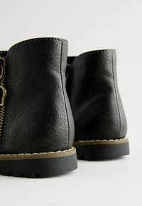 Paire de bottines en cuir noir avec surpiqûres marron clair et semelles en caoutchouc noir, vues de dos sur un fond blanc.
