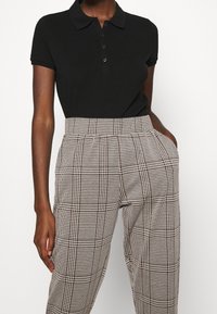 Černé polo triko s krátkými rukávy a zapínáním na knoflíky, v kombinaci s vysokým pasem kostkovaných kalhot v černobílém vzoru.