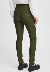 Jeans skinny de tiro alto en color verde oliva con una textura suave, bolsillos traseros y detalles en botón plateado, combinados con zapatos negros sin cordones.