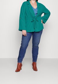 Jadegrön kavaj med skärp och vida ärmar, över en blå mönstrad topp. Kombinerad med smala jeans och bruna stövletter med klack.