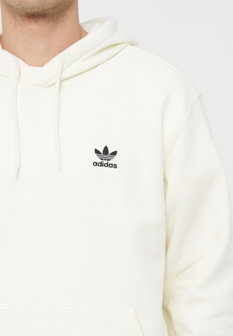 Osoba nosząca białą bluzę Adidas z kapturem z czarnym logo trójlistka na piersi i sznureczkami zwisającymi z kaptura.