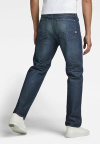 Dark blue denim jeans with a straight cut, featuring two back pockets and subtle stitching details. Paired with white sneakers.