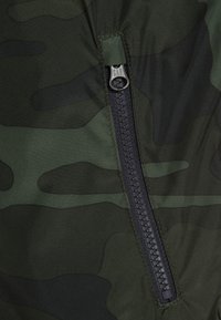 Gros plan sur une veste camouflage en tissu vert foncé à finition brillante, avec une fermeture éclair noire portant un logo en relief.
