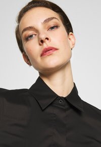 Black collared shirt with a button closure detail, featuring a smooth texture and fitted design. The collar is slightly structured.