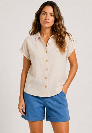 Femme aux longs cheveux ondulés portant une chemise beige à manches courtes boutonnée et un short bleu à revers, debout avec une main dans la poche.