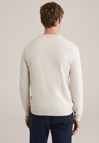 Homme aux cheveux courts et bouclés, vêtu d'un pull crème à manches longues et d'un pantalon foncé, vu de dos sur un fond uni.