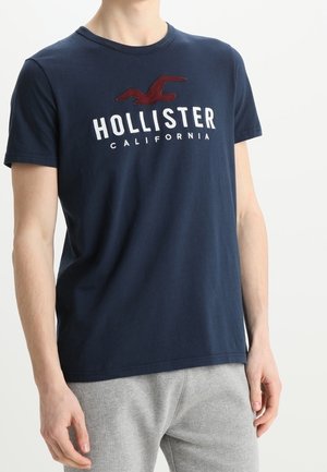 Persoon draagt een marineblauw Hollister California T-shirt en lichtgrijze broek, staand tegen een effen lichte achtergrond.