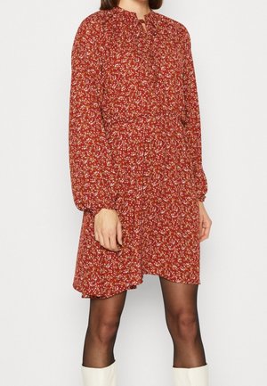 Mujer que lleva un vestido largo de manga larga con estampado floral rojo, combinado con medias negras transparentes y botas blancas hasta la rodilla, sobre un fondo liso.