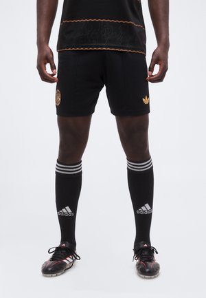 Mannelijke atleet met zwarte Adidas-shorts, zwarte Adidas kniehoge sokken met witte strepen en zwarte voetbalschoenen met rode accenten.