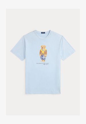 Gaiši zila kokvilnas T-krekls ar druku, kurā attēlots lācis brūnā jakā, dzeltenā džemperī un zilās biksēs. Zem tā teksts "Polo Bear by Ralph Lauren".