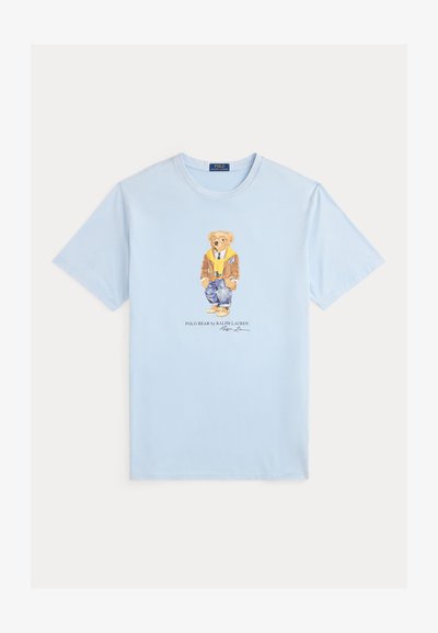 Gaiši zila kokvilnas T-krekls ar druku, kurā attēlots lācis brūnā jakā, dzeltenā džemperī un zilās biksēs. Zem tā teksts "Polo Bear by Ralph Lauren".