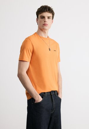 Jaunas vyras dėvi oranžinį BOSS marškinėlį ir tamsius džinsus, stovi kišenėse rankas įdėjęs priešais baltą, paprastą foną.