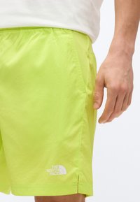 Άνδρας που φοράει λαμπερά πράσινα λάιμ σορτς με το λογότυπο The North Face στο αριστερό πόδι, στέκεται μπροστά σε ανοιχτό φόντο.