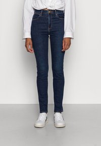 Mörkblå skinny jeans med hög midja, fem fickor och guldhardware, kombinerat med vita sneakers på ett ljust grått golv.