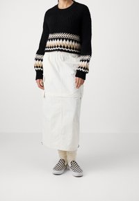 Czarny sweter dziany z wzorzystym beżowo-białym poziomym pasem, zestawiony z białą spódnicą, białymi skarpetkami i czarno-białymi wsuwanymi butami w kratę.