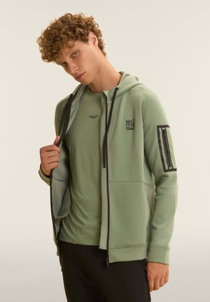 Junger Mann mit lockigem Haar, der eine hellgrüne Kapuzenjacke mit Reißverschluss und ein passendes T-Shirt trägt und posiert, während er mit einer Hand die Kapuzenjacke geöffnet hält.