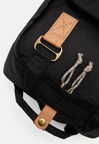 Black fabric backpack with tan leather accents. Features a metal D-ring and decorative stitching. Includes decorative cord pulls.