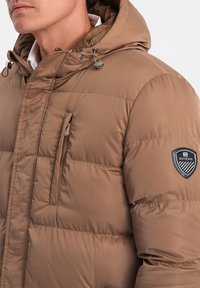 Giacca imbottita marrone con cappuccio, dotata di una tasca con zip e un patch logo sulla manica. Materiale liscio e leggero con una leggera lucentezza.