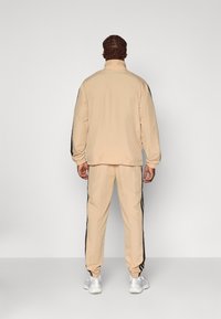 Survêtement de sport beige comprenant une veste zippée avec un col haut, un pantalon assorti et des bandes latérales noires contrastantes. Texture lisse.