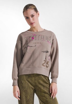 Vrouw met naar achteren gekamd haar draagt een beige sweater met "ATTITUDE" en schetsen, gecombineerd met olijfgroene broek, staand tegen een witte achtergrond.