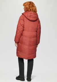 Rode lange gewatteerde jas met een capuchon, voorzien van een gewatteerd ontwerp, elastische manchetten en een tailleband, gecombineerd met zwarte broek en stevige zwarte laarzen.