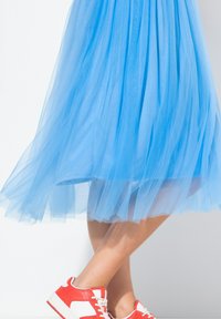 Gonna in tulle azzurro chiaro con texture a strati, abbinata a sneakers bianche e rosse. Focus sul design fluido e sul tessuto morbido.