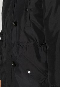 Černá vyteplená bunda s knoflíkovým zapínáním, bočními kapsami a hladkým povrchem. Tkanina vypadá odolně s matným vzhledem.