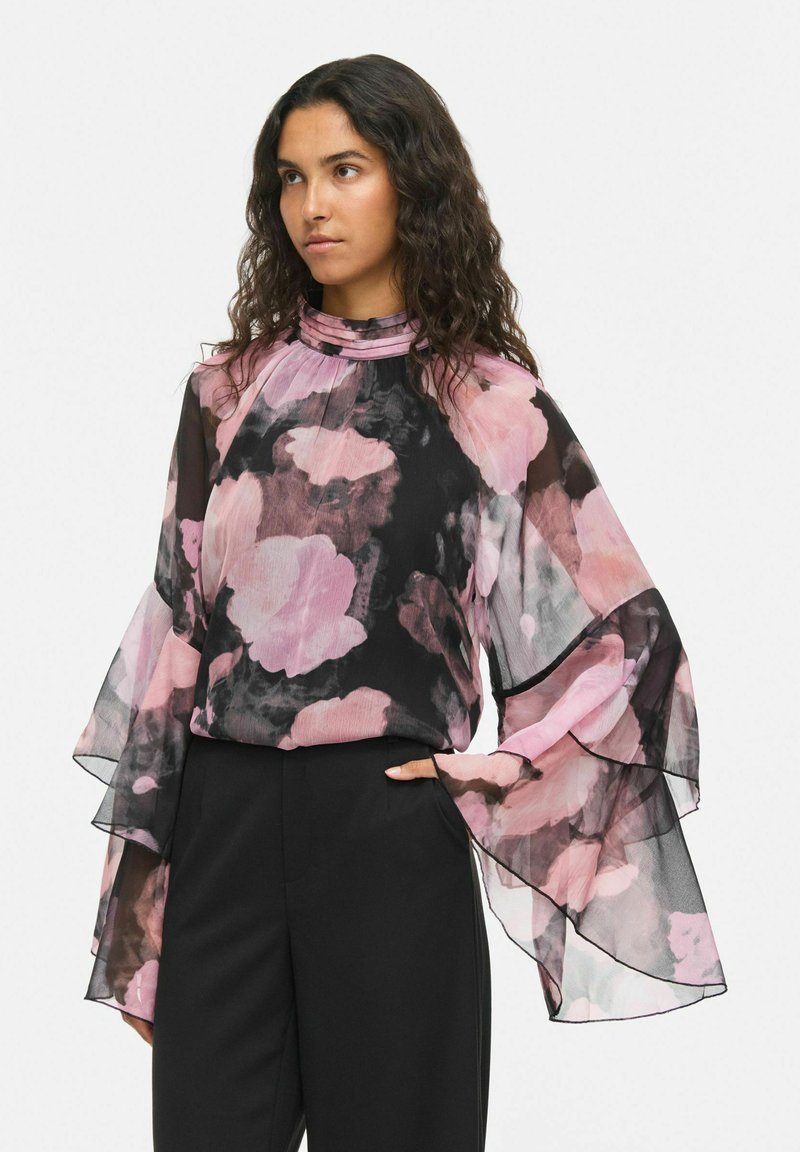 Femme aux longs cheveux bouclés portant un chemisier transparent noir et rose à motif floral avec des manches cloche superposées et un pantalon noir.