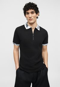 Νεαρός άνδρας με σγουρά μαλλιά που φοράει μαύρο πουκάμισο με κοντό μανίκι και λευκό γιακά και μανσέτες με μαύρο κείμενο, με τα χέρια στις τσέπες, ουδέτερο φόντο.