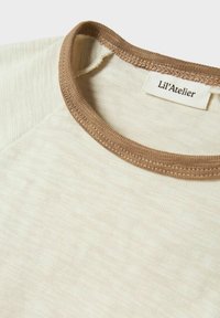 Cremfarbendes Hemd mit braunem geripptem Kragen, das eine weiche Textur aufweist und ein Etikett mit der Aufschrift "Lil'Atelier" trägt.