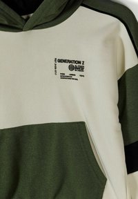 Sweat à capuche avec blocs de couleurs crème et olive, poche avant, et inscription noire "Generation Z" ainsi que les noms des villes Paris, Londres, Tokyo.
