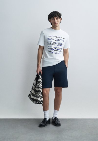 Λευκό βαμβακερό T-shirt με μπλε κυματιστά σχέδια, σε συνδυασμό με σκούρο μπλε σορτς. Συνδυάζεται με μαύρη και λευκή τσάντα tote με ζιγκ-ζαγκ σχέδιο. Φοριούνται μαύρα παπούτσια.