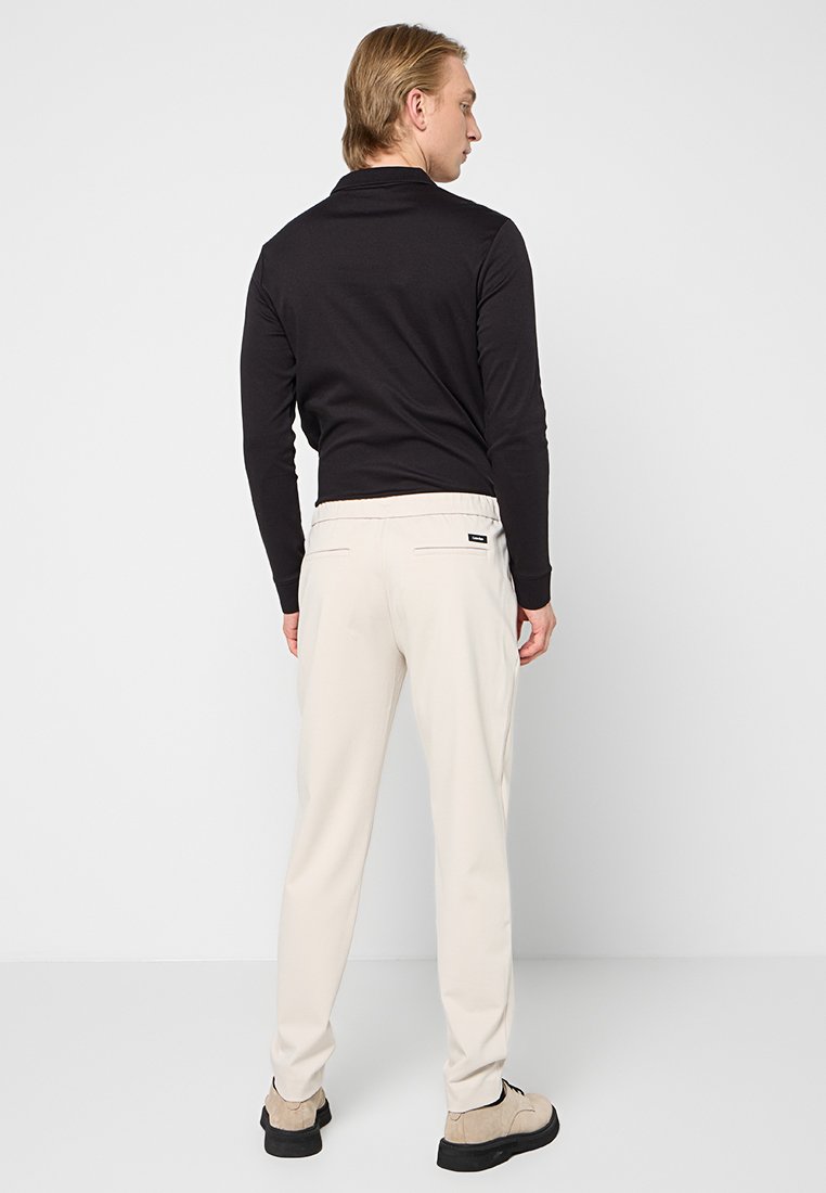 Schwarzes Langarm-Polo-Shirt kombiniert mit beigen Hosen, mit schlanker Passform und elastischem Bund. Die Schuhe sind hell mit dunklen Sohlen.