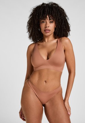 Frau mit lockigem Haar, die einen hautfarbenen nahtlosen BH und Unterwäsche trägt, steht vor einem einfarbigen hellen Hintergrund und schaut nach vorne.