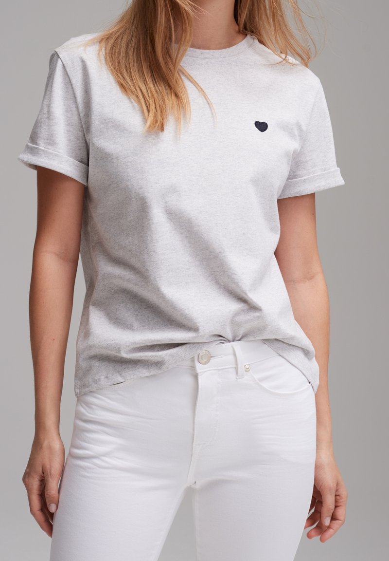 Femme portant un t-shirt gris clair à manches courtes avec un petit cœur noir sur la poitrine gauche et un pantalon blanc ajusté sur fond neutre.