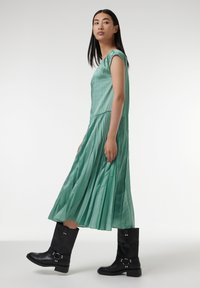 Mintgrünes, plissiertes Kleid mit kurzen Ärmeln und taillierter Passform, kombiniert mit schwarzen kniehohen Stiefeln mit metallischen Schnallenakzenten.
