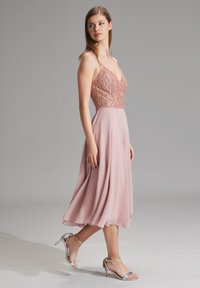 Knielang, A-lijn jurk in lichtroze, met een bewerkte top, dunne bandjes en een vloeiende chiffon rok, gedragen met zilveren sandalen met hakken.