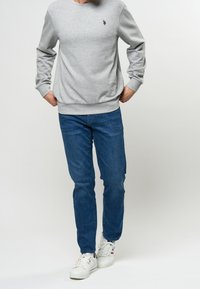 Sudadera gris con cuello redondo y puños acanalados, combinada con jeans de color azul oscuro. Zapatillas blancas con cordones completan el atuendo.