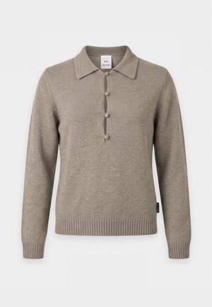 Kvinna som bär en taupefärgad pikétröja med långa ärmar, svart läderkjol, vit skjorta knuten i midjan, svarta snörkängor till knäna och bär en mörkgrön axelväska.